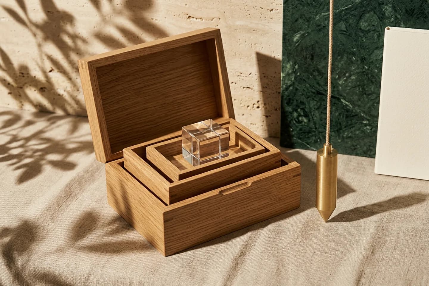 Three nested oak boxes on linen, the smallest holding a glass cube, with a brass plumb line hanging beside them.