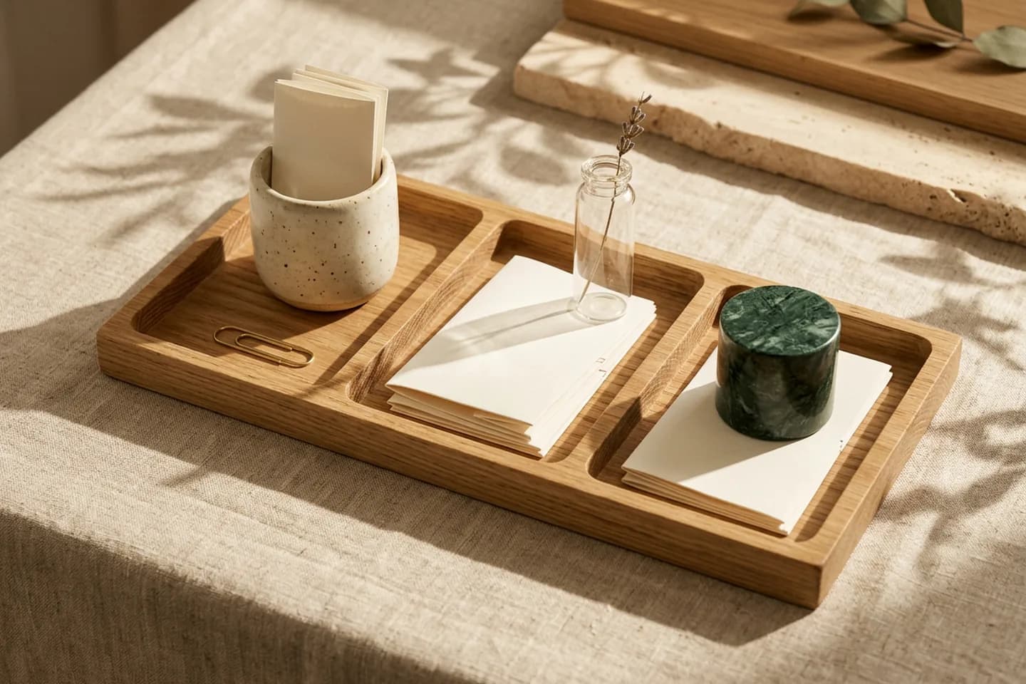 Uma bandeja de carvalho com três compartimentos contendo folhas de papel marfim em branco organizadas, cada uma ao lado de um pequeno vaso de vidro, cerâmica ou mármore, sobre linho sob luz quente do sol.