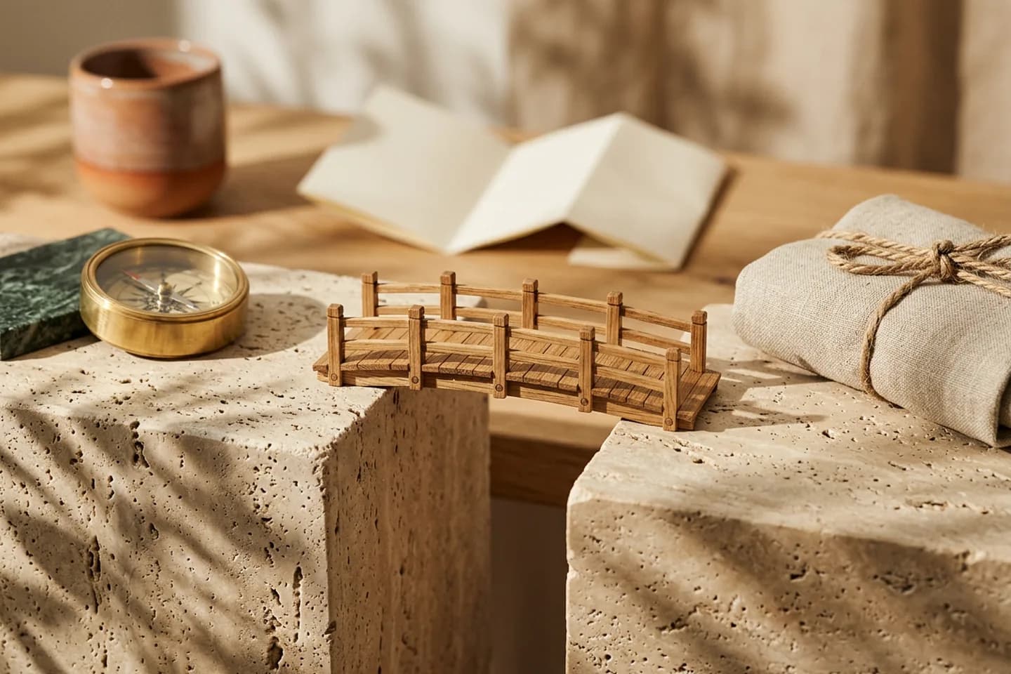 An oak bridge model connecting two travertine blocks, with a brass compass on one side and a rope-tied linen bundle on the other.