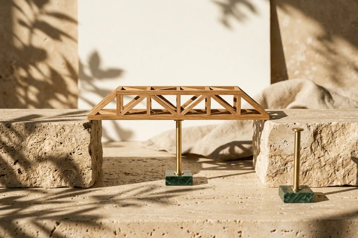 A small oak bridge model resting on a single slim brass pier between two travertine blocks, with a spare brass pier standing beside it in warm sunlight.