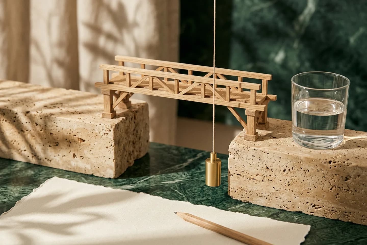 Modelo de ponte em carvalho cruzando o vão entre dois blocos de travertino, com um prumo de latão pendurado ao lado de um dos pilares sob luz quente do sol.